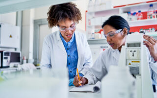Two scientists standing in a lab and writing data in a notebook