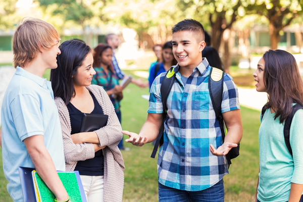 Teenage Hispanic male college student is telling story to group of friends outdoors on school campus. Male and female students are standing in a group, discussing something after class. They are wearing casual clothing and carrying school books.