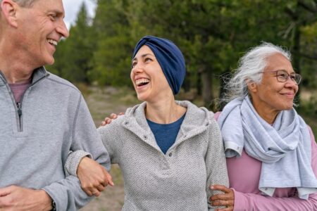 A young mixed race woman with cancer laughs while spending time outdoors with her supportive and loving parents. Their arms are linked and they are walking in a forested area. All of the individuals are smiling and feeling positive about the future.